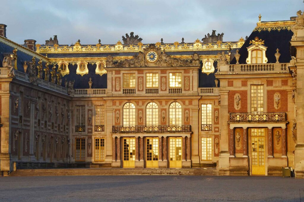 The exterior of Airelles Château de Versailles with the sunset reflecting on its windows.