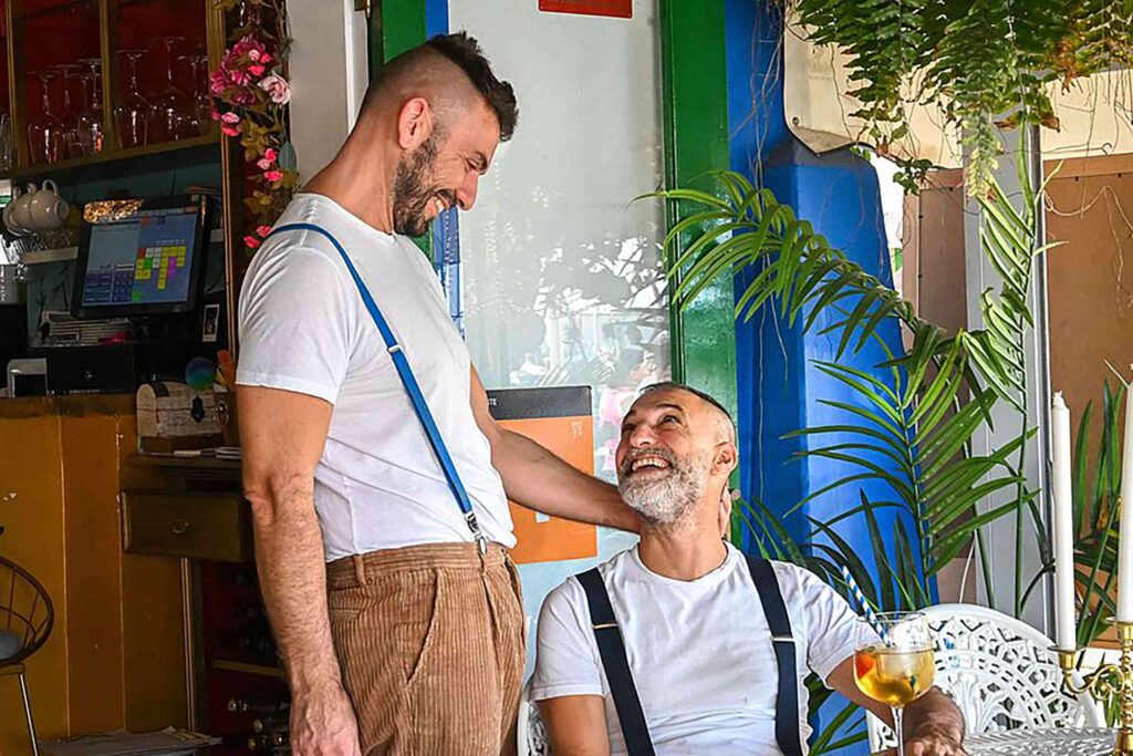 Mauro and Carmello, partners in life and work at Mi Vida restaurant in Puerto de Mogán look and smile at each other outside their restaurant. Both are wearing white t-shirts and braces, one has a brown beard and the other a grey beard