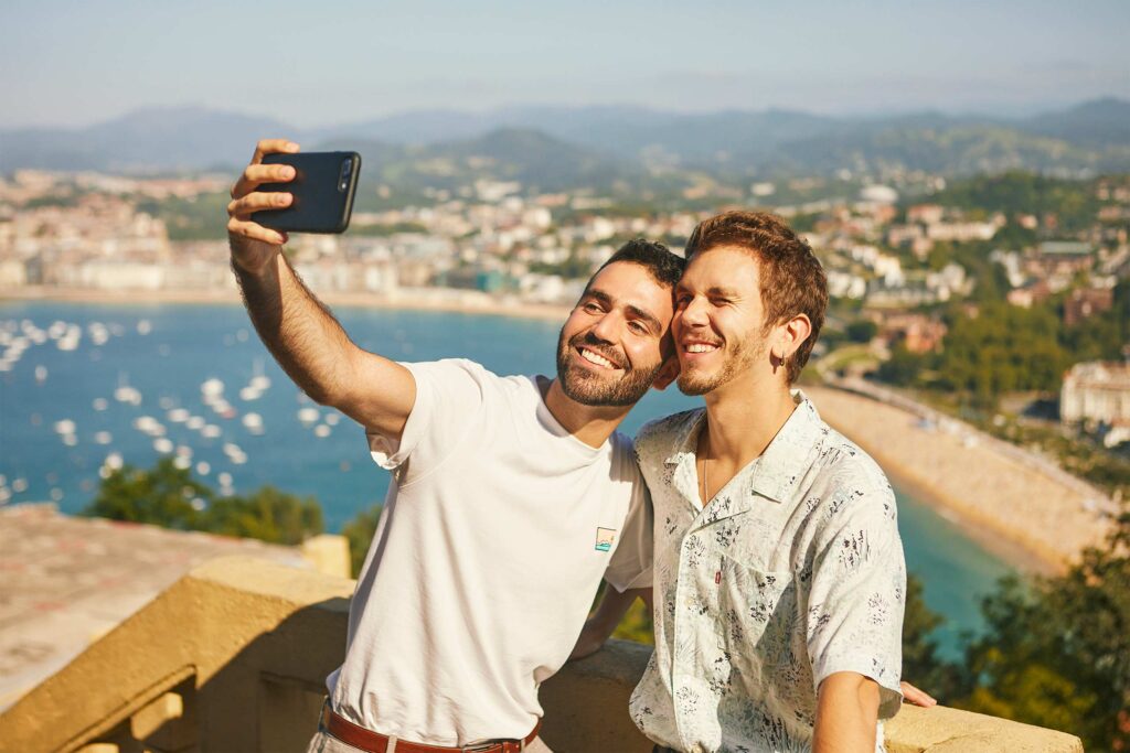 A gay couple take a selfie from a viewpoint in San Sebastian with a beach in the background.