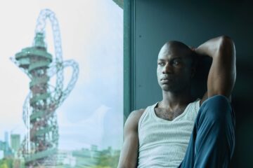 Black model in a white vest and his arm up sits by a window with the view of the ArcelorMittal Orbit in London