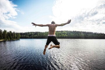 Topless man with long black shorts and pink underwear jump dives into the sea water in the archipelago around Stockholm. The background is a horizon of pine trees.