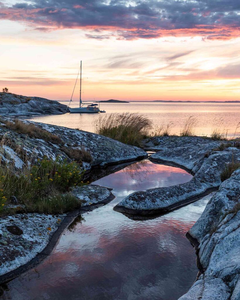 View of the Stockholm Archipelago at sunset over a rocky islet. It's golden hour and their are rockpools full of water reflecting the red sky. A sailboat is moored on the island.