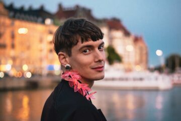 Queer, non-binary person with a pink feather earring looks happily to camera with Stockholm in the background