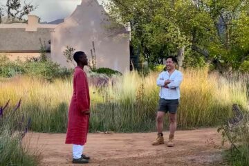 South African man in a long ochre robe talks to male Asian tourist in a white shirt, shorts and tan boots in the gardens of on a healing farm hotel in South Africa