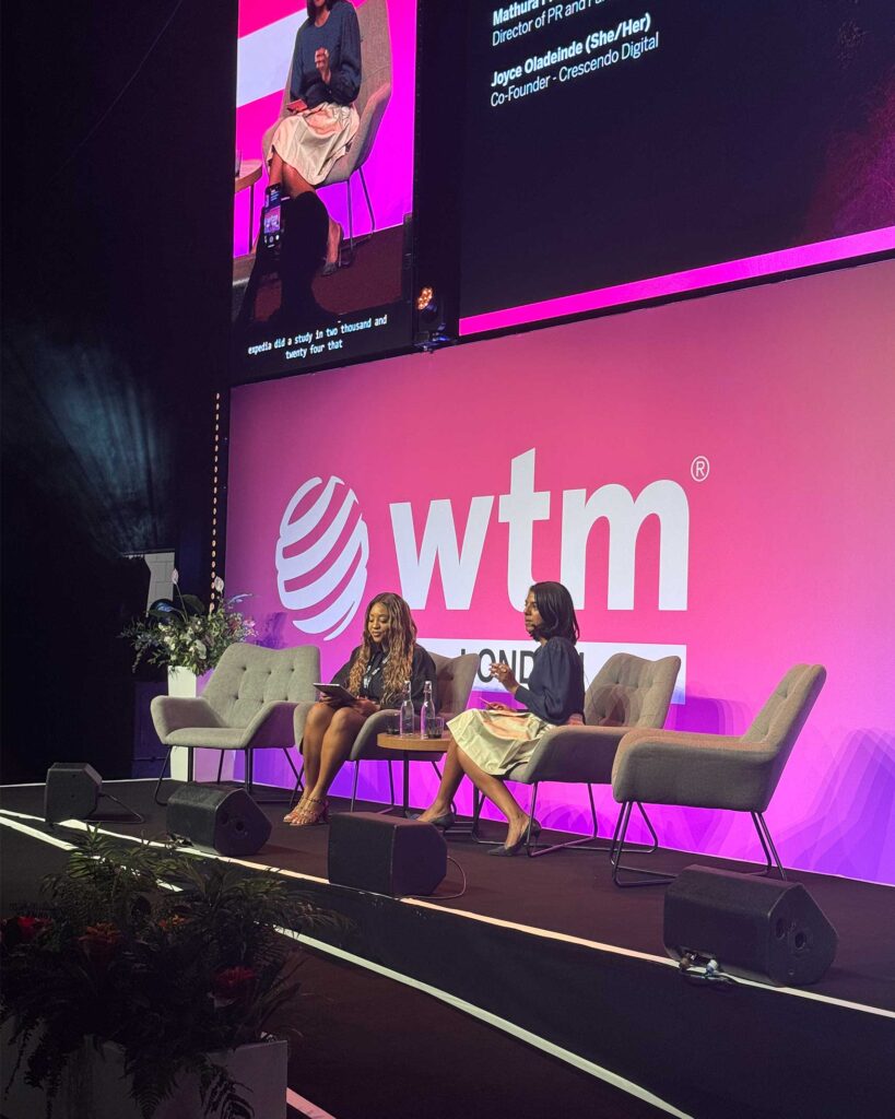 Two women in conversation on stage at WTM London’s Orange Theatre. One speaks while holding a microphone, and the other listens with a smile, both seated in grey chairs under bright lighting.