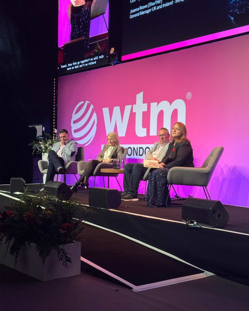 Four panellists seated on stage, engaged in discussion beneath a large screen showing a close-up of the session speaker. The event logo is brightly displayed behind them.